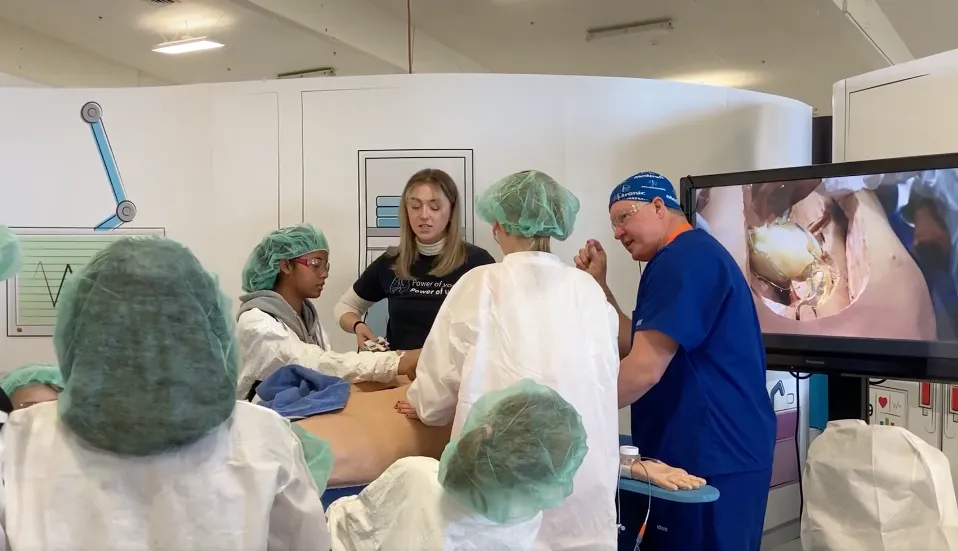 Small kids wearing white lab coats and green hairnets gather around a "patient"