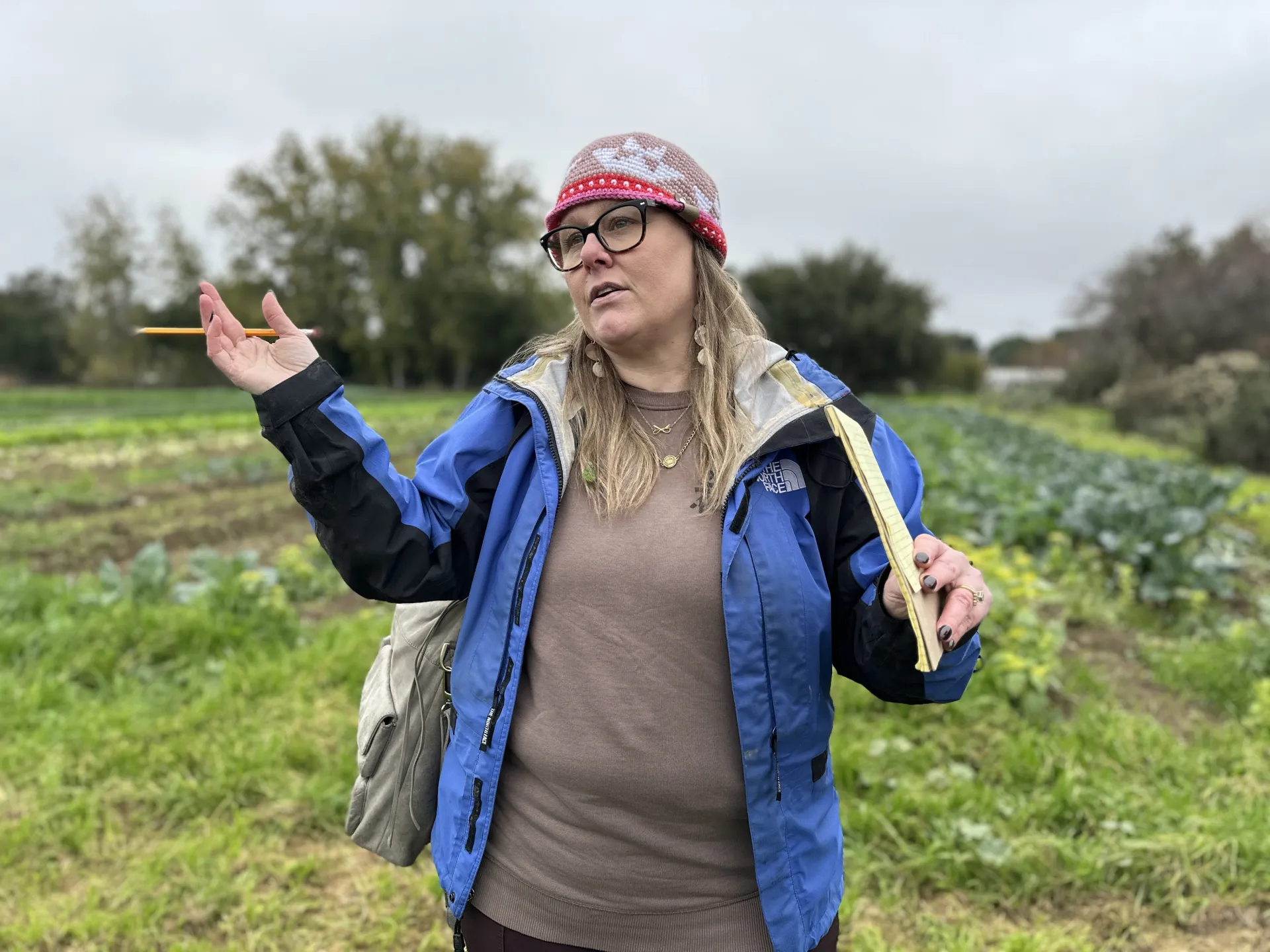 A woman standing in a green field, wearing a blue jacket and a cap, gesturing with her hands