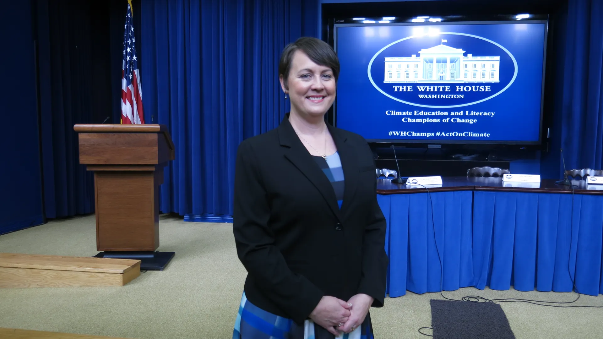 Woman wearing a black blazer smiles as she stands in a White House meeting room with a podium and table in the background