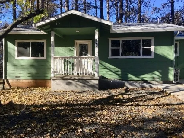 An intact green house with no vegetation in front. Several singed trees behind the house.