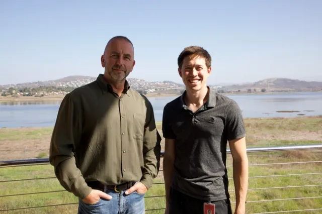 Two men stand outdoors in a park with a reservoir behind them.