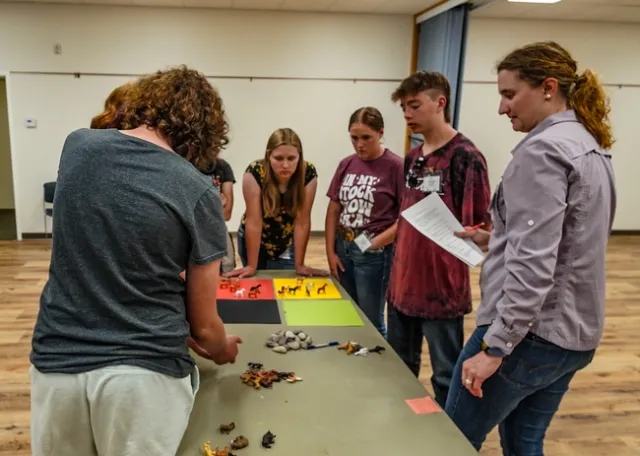 Snell holds a sheet of paper and pen as she observes teenagers placing toy horses on black, orange, yellow and green squares of paper.
