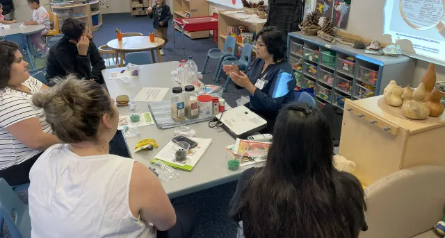 EFNEP Community Educator at a table teaching an ESBA lesson to 4 adults in an elementary school classroom.