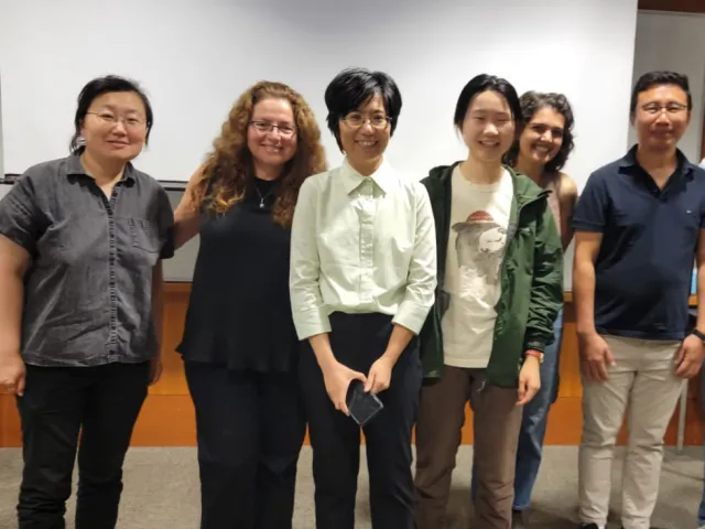 Six men and women stand in a row, smiling in a lecture hall or classroom setting