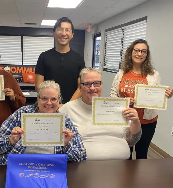Burkes, seated, hold up their EFNEP course completion certificates as Danny Vang stands smiling behind them