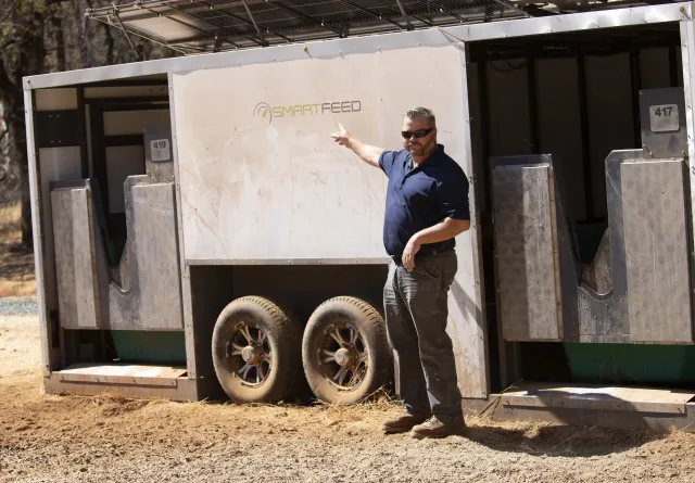 Man in blue polo shirt and sunglasses points to a SmartFeeder machine, which looks like a trailer with openings where cows put their heads in