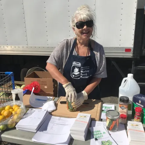 woman standing in front of a delivery truck and behind a table with lemons, papers, jarred vegetables, vinegar, salt and spices