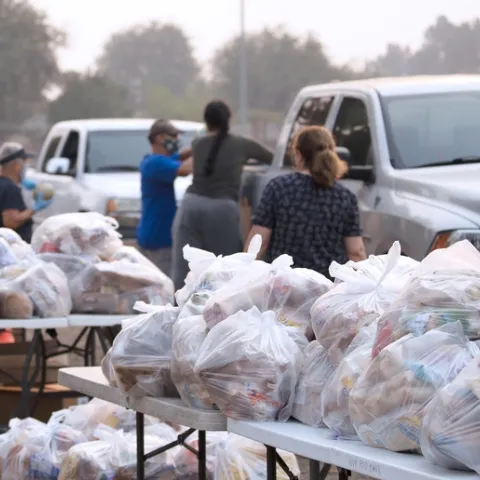 In the foreground are tables with mounds of white plastic bags filled with food items. In the background are volunteers wearing facemasks placing bags of food in the back of a silver pickup and a white pickup.