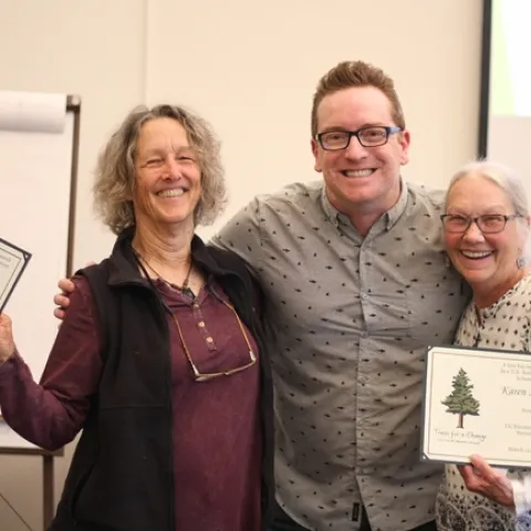 Three adults with big smiles and two of them are holding certificates in their hand.
