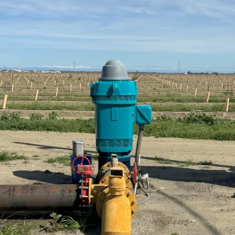 A blue and yellow well pump is shown at the edge of a field of saplings.