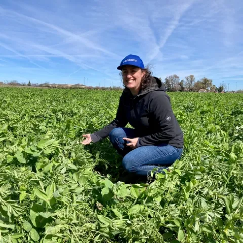 Sarah is wearing a blue UC ANR hat and crouching in a luscious green field under a blue sky.