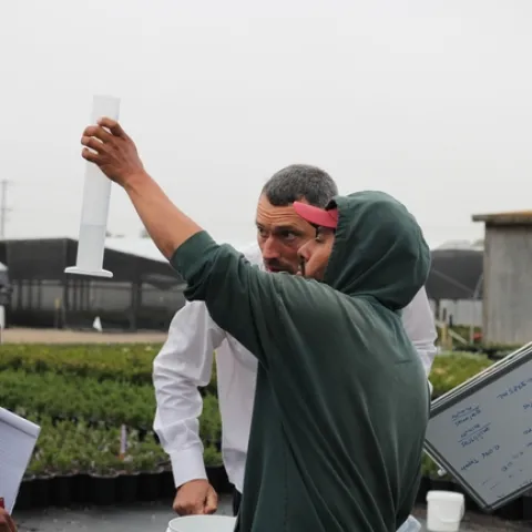 Two men stand side by side. One holds up a beaker with water and the other looks closely at it to measure how much water is captured.