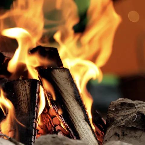 Close-up of a campfire with burning logs surrounded by a circle of rocks. In the blurred background, a green and orange tent and a picnic basket are visible.