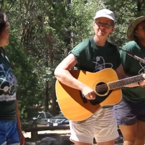 Marianne strums guitar surrounded by 3 other women wearing matching dark green t-shirts
