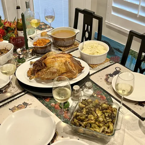 A dining table with stuffing, sweet potatoes, gravy, a roast turkey, mashed potatoes, Brussels sprouts, green bean casserole, and 5 glasses of wine.