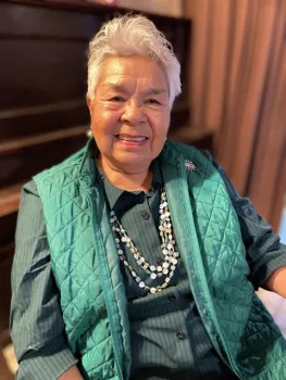 An older woman seated and smiling and wearing a light green vest
