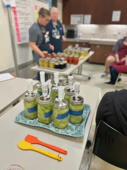 Jars with fermentation lids filled with green cabbage in forefront and two volunteers examine jarred products in the background. 