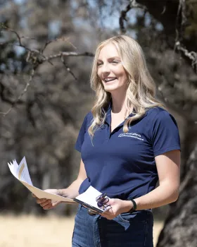 Andrea Warner, wearing a blue polo, holds papers and gestures while she speaks in a grove of trees